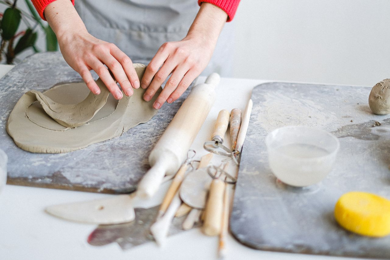 Unrecognizable female master standing at table with set of tools and wooden boards while working with cut piece of clay